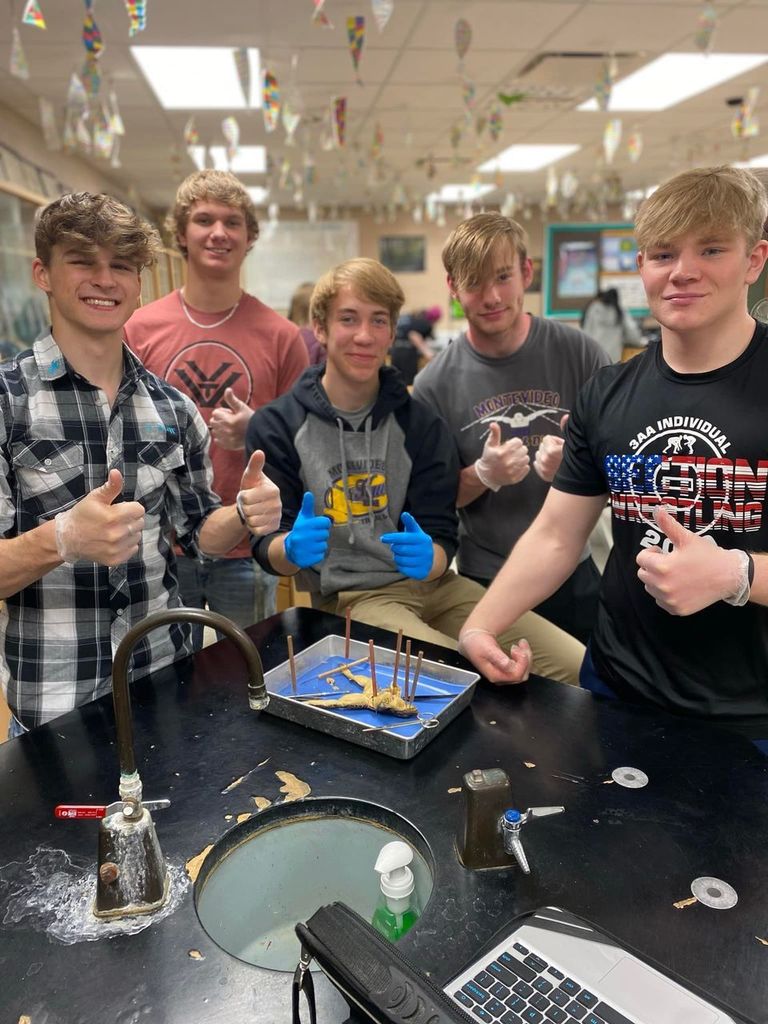 Students in biology classroom holding tray with dissected frog and smiling 